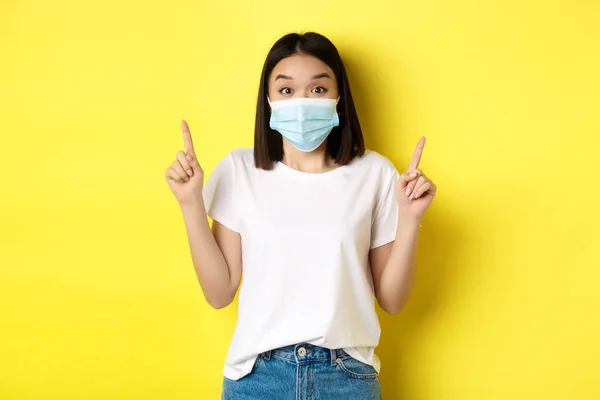 Covid-19, pandemiskt och socialt distanserande koncept. Ung asiatisk kvinna i vit t-shirt och medicinsk mask från coronavirus, pekar finger upp, visar specialerbjudande — Stockfoto