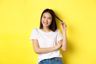 Kendinden emin ve şık Asyalı kadın göğsünde çapraz kollar ve gülümsüyor, sarı arka planda duruyor.