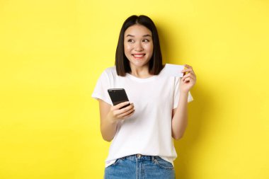 E-ticaret ve online alışveriş konsepti. Neşeli Asyalı kız internetten ödeme yapıyor, elinde akıllı telefon ve plastik kredi kartı, gülümsüyor ve sola bakıyor, sarı arka plan