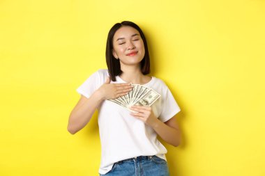 Güzel Asyalı kadın parayı sever, dolarları sever ve gülümser lütfen, sarı arka planda durur.