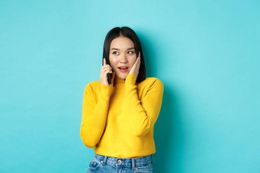 Cep telefonuyla konuşan bir kadın ilginç görünüyor. Arama sırasında ilginç bir teklif alıyor. Mavi arka planda duruyor.