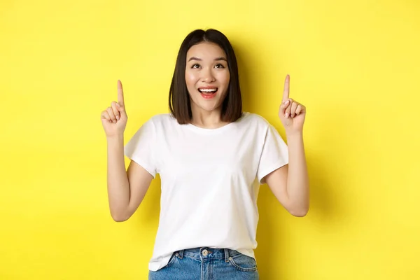 Concepto de belleza y moda. Hermosa mujer asiática en camiseta blanca apuntando con los dedos hacia arriba, de pie sobre fondo amarillo —  Fotos de Stock