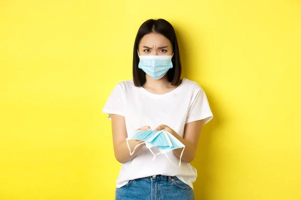 Coronavirus, καραντίνα και ιατρική έννοια. Θυμωμένη Ασιάτισσα σου δίνει ιατρική μάσκα σε εσωτερικούς χώρους, συνοφρυωμένη αναστατωμένη, στέκεται πάνω από κίτρινο φόντο — Φωτογραφία Αρχείου