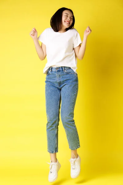 Tam boy, kaygısız Asyalı kadının zıplaması ve dans etmesi, eğlenmesi, kot pantolon ve beyaz tişörtle sarı arka planda poz vermesi. — Stok fotoğraf