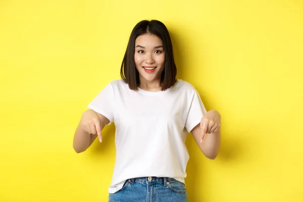 Concetto di bellezza e moda. Bella donna asiatica in t-shirt bianca puntando il dito verso il basso, dimostrare logo in piedi su sfondo giallo — Foto Stock