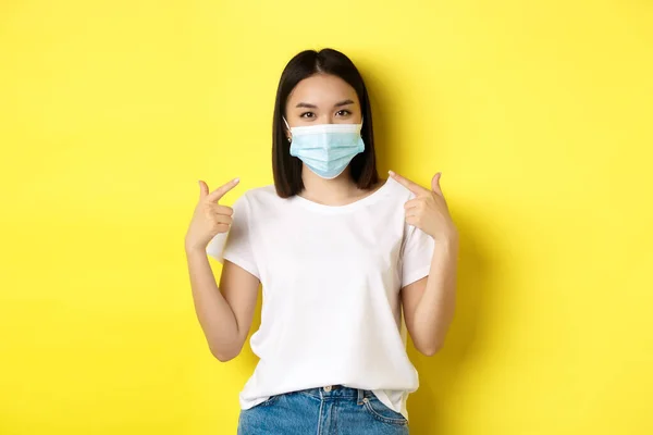 Covid-19, karantän och socialt distanserande koncept. Ung asiatisk kvinna i vit t-shirt, pekar på sin medicinska mask från coronavirus, gul bakgrund — Stockfoto