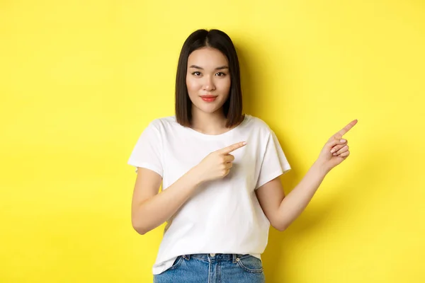 아름다움과 패션 개념. 흰색 티셔츠를 가리키는 손가락을 오른쪽으로 하고 노란색 배경 위에 서 있는 아름다운 동양 여성이 로고를 보여 주고 있다 — 스톡 사진