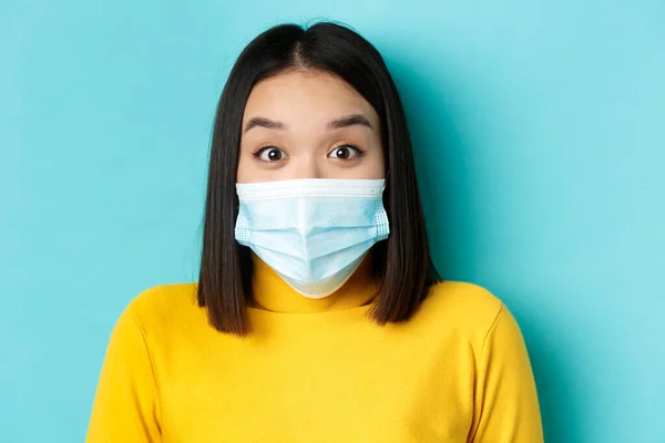 Covid-19, socialt avståndstagande och pandemikoncept. Närbild av förvånad asiatisk kvinna i medicinsk mask, höja ögonbryn och titta förvånad över kameran, blå bakgrund — Stockfoto
