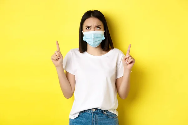 Covid-19, conceito de pandemia e distanciamento social. Desapontado menina asiática em máscara médica, franzindo a testa chateado e apontando os dedos para o logotipo, de pé sobre fundo amarelo — Fotografia de Stock