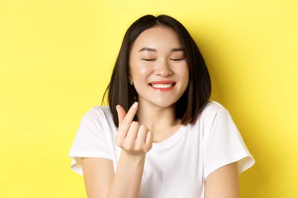 Dia dos Namorados e conceito de mulheres. Close up de menina asiática bonita em t-shirt branca, sorrindo e mostrando o coração do dedo, eu te amo gesto, de pé sobre fundo amarelo — Fotografia de Stock