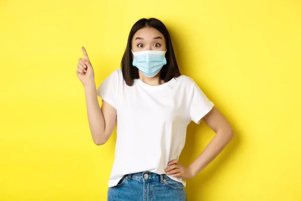 Ковид, здравоохранение и пандемия. Азиатская модель в медицинской маске и белой футболке указывая пальцем на логотип верхнего левого угла, показывая продвижение, желтый фон — стоковое фото
