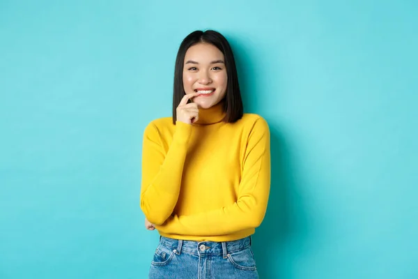 Image de belle femme asiatique en tenue élégante, lèvres touchantes et souriant à la caméra avec un visage heureux, debout sur fond bleu — Photo