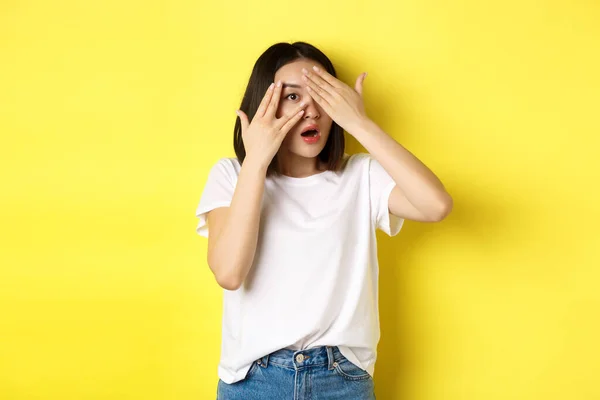 Kíváncsivá tett ázsiai lány vár meglepetés, kukucskál az ujjain keresztül a szemét, mosolygós boldog, álló felett sárga háttér — Stock Fotó
