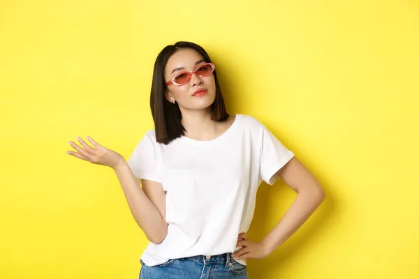 Concepto de moda y estilo de vida. Mujer asiática atrevida y confiada en gafas de sol de moda con aspecto seguro de sí mismo en la cámara, de pie sobre el fondo amarillo —  Fotos de Stock