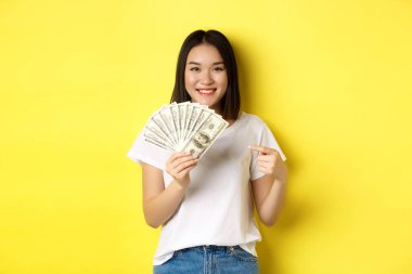 Genç Asyalı kadın gülümsüyor, ödül parasını gösteriyor, dolarları işaret ediyor, sarı arka planda duruyor.