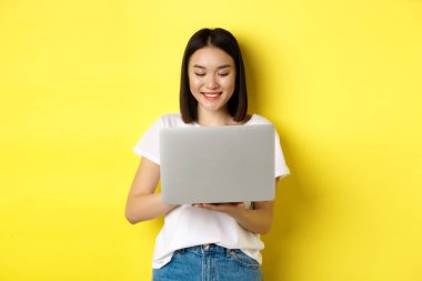 Güzel Asyalı kadın dizüstü bilgisayarda çalışıyor, ekranda mutlu görünüyor, sarı arka planda duruyor.