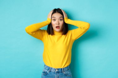 Şaşırmış Asyalı kadın nutku tutulmuş, hayrete düşmüş ve el ele tutuşmuş, mavi arka planda dikilen büyük haberi duyuyor.