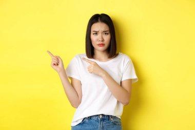 Güzellik ve moda konsepti. Beyaz tişörtlü güzel Asyalı kadın, sarı arka planda durup, parmaklarını sola gösteriyor.