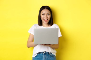Neşeli Asyalı kadın dizüstü bilgisayarda çalışıyor ve gülümsüyor, beyaz tişört ve kot pantolonla sarı arka planda duruyor.