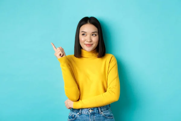 Conceito de compras. Elegante asiático modelo feminino em camisola amarela, sorrindo e apontando dedo esquerdo, mostrando propaganda com rosto satisfeito, de pé sobre fundo azul — Fotografia de Stock
