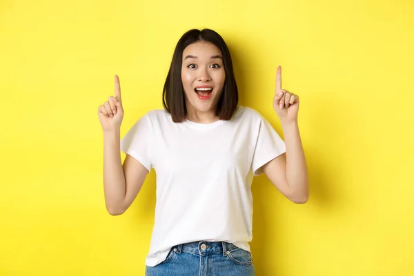 Concepto de belleza y moda. Hermosa mujer asiática en camiseta blanca apuntando con los dedos hacia arriba, de pie sobre fondo amarillo —  Fotos de Stock