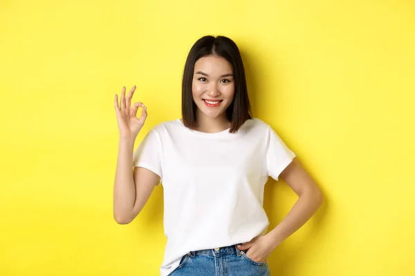 Kendine güvenen Asyalı kadın gülümsüyor ve tamam işareti gösteriyor. Onaylıyor ve iyi bir teklif sunuyor. Sarı arka planda beyaz tişörtle duruyor. — Stok fotoğraf