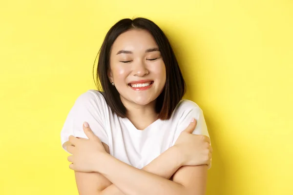 Romantik Asyalı kızın kendine sarılıp hayal kurması, gözlerini kapatması ve gülümsemesi, sarı arka planda durduğunu hayal ederken. — Stok fotoğraf