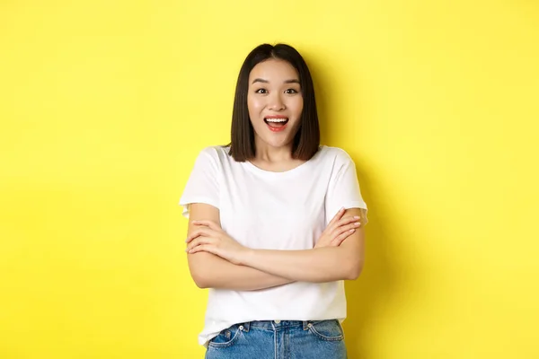 Concepto de belleza y moda. Mujer asiática feliz en camiseta blanca, brazos cruzados en el pecho, mirando asombrado a la cámara, de pie sobre fondo amarillo —  Fotos de Stock