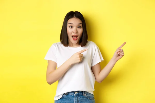 Concepto de belleza y moda. Hermosa mujer asiática en camiseta blanca señalando los dedos a la derecha, demostrar el logotipo de pie sobre el fondo amarillo — Foto de Stock