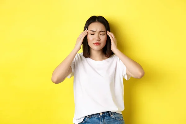 Portret van een Aziatische vrouw, ogen dicht en frons van migraine, aanrakend hoofd, duizelig van hoofdpijn, staand over een gele achtergrond — Stockfoto