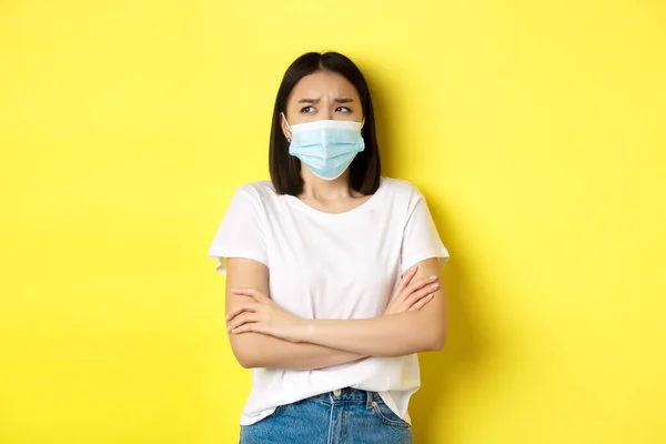 Covid, gezondheidszorg en pandemie concept. Aziatische vrouw in wit t-shirt en medisch masker kruis armen op de borst en op zoek omhoog pensive en bezorgd — Stockfoto