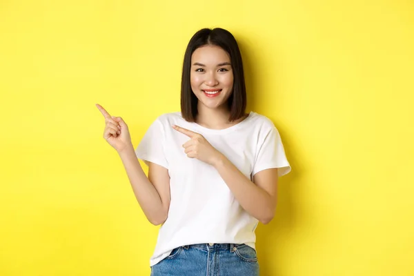 Concepto de belleza y moda. Hermosa mujer asiática en camiseta blanca señalando los dedos a la izquierda, de pie sobre fondo amarillo —  Fotos de Stock