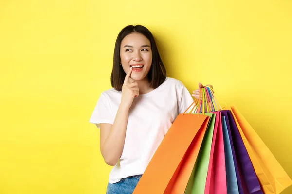 Penzitivní mladá žena se zaujatě usmívá, ukazuje nákupní tašky, přemýšlí o koupi něčeho, stojí nad žlutým pozadím — Stock fotografie