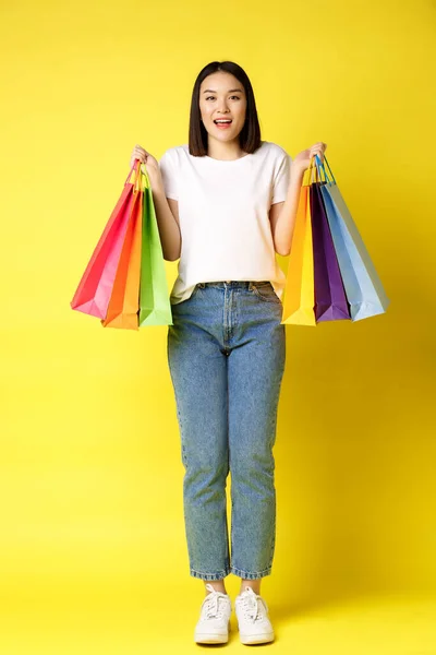 Plná velikost portrét krásné asijské dívky jít nakupovat, drží papírové tašky z obchodů a usmívá se, stojí v džínách a bílé tričko přes žluté pozadí — Stock fotografie