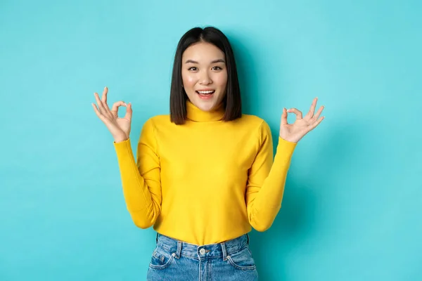 Alegre asiático modelo feminino mostrando ok gestos, sorrindo e olhando impressionado, produto de louvor, de pé sobre fundo azul — Fotografia de Stock