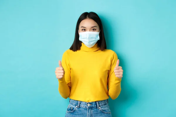Covid-19, socialt avståndstagande och pandemikoncept. Stödjande asiatisk kvinna rekommenderar befordran, bär medicinsk mask och visar tummen upp, står över blå bakgrund — Stockfoto