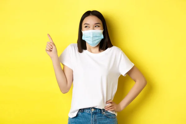 Covid, sjukvård och pandemi koncept. Asiatisk kvinna i medicinsk mask och vit t-shirt pekar finger på övre vänstra hörnet logotyp, visar befordran, gul bakgrund — Stockfoto