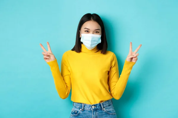 Covid-19, concetto di distanza sociale e pandemia. Kawaii donna asiatica che mostra segni di pace e sorridente felice, indossando maschera medica dalla malattia coronavirus, in piedi su sfondo blu — Foto Stock