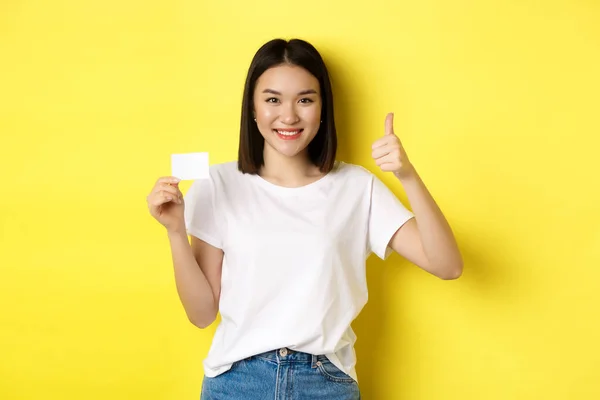 Plastik kredi kartı gösteren, jest yapan, onaylayan ve tavsiye eden, kameraya gülümseyen, sarı arka planlı sıradan beyaz tişörtlü genç Asyalı kadın. — Stok fotoğraf