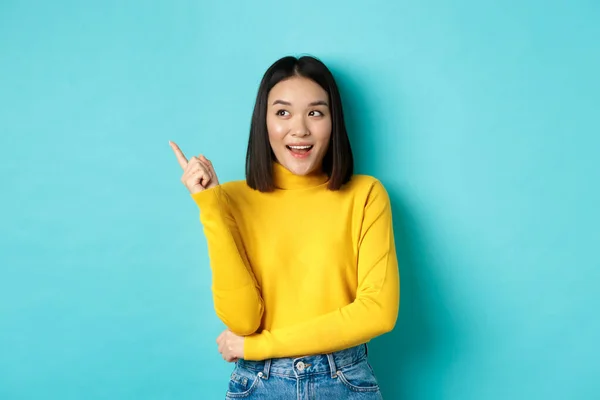 Köpkoncept. Porträtt av attraktiv koreansk flicka i gul tröja, visar befordran erbjudande på kopiera utrymme, pekar och tittar vänster med nöjda leende, blå bakgrund — Stockfoto