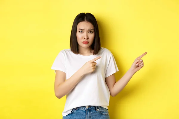 Concepto de belleza y moda. Hermosa mujer asiática en camiseta blanca señalando los dedos a la derecha, demostrar el logotipo de pie sobre el fondo amarillo — Foto de Stock