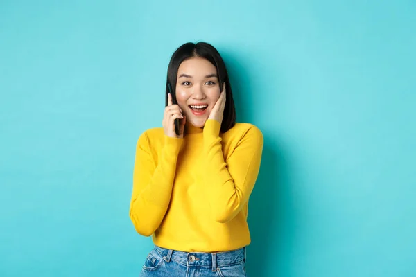 행복 한 이시아 여성은 스마트폰으로 말하면서 웃으면서 전화 통화를 하면서 푸른 배경 위에 서서 제공되는 제안을 받는다 — 스톡 사진