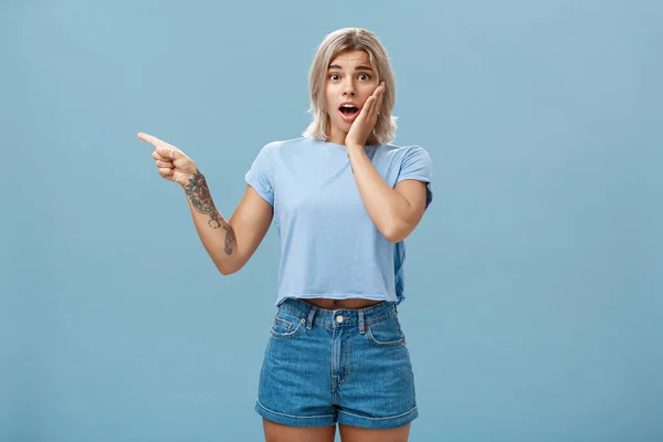 Pas-up záběr ustaraný překvapený emotivní blondýny v tričku říká gosh otevření úst od šoku drží dlaň na tváři při pohledu s empatií, zatímco ukazuje vlevo s vytetovanou rukou přes modrou zeď — Stock fotografie