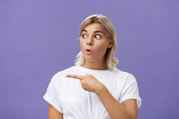 Primer plano de intrigado interesado atractivo adulto rubia hembra en blanco camiseta plegable labios mirando fijamente y señalando a la izquierda con expresión curiosa y emocionada sobre fondo púrpura — Foto de Stock