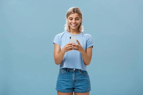 Flicka som har roligt convesation, talar med vän via videomeddelanden i trådlösa hörlurar håller smartphone i båda händerna ler glatt på enheten skärmen underhålls och roas — Stockfoto