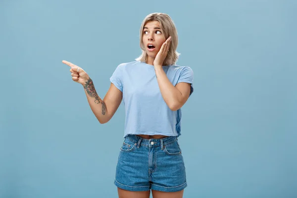 Studio shot van nerveus geschokt goed uitziende jonge vrouw met mooi haar en getatoeëerde arm hijgend wijzend en kijkend links met zorg hand op hand houden op de wang poseren over blauwe achtergrond — Stockfoto