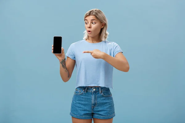 Você deve baixar aplicativo. Retrato de surpreendido chocado menina loira atraente na moda verão t-shirt segurando smartphone ofegante de surpresa apontando para tela gadget posando sobre fundo azul — Fotografia de Stock