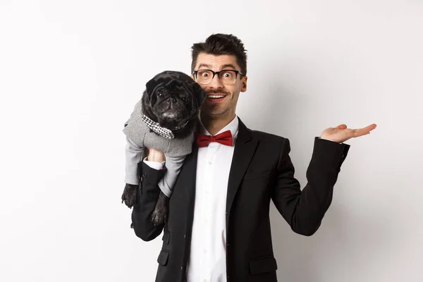 Stilig ung man introducerar sällskapsdjur produkt på hand, håller söt svart hund på axeln och ler, visar något på vit bakgrund — Stockfoto