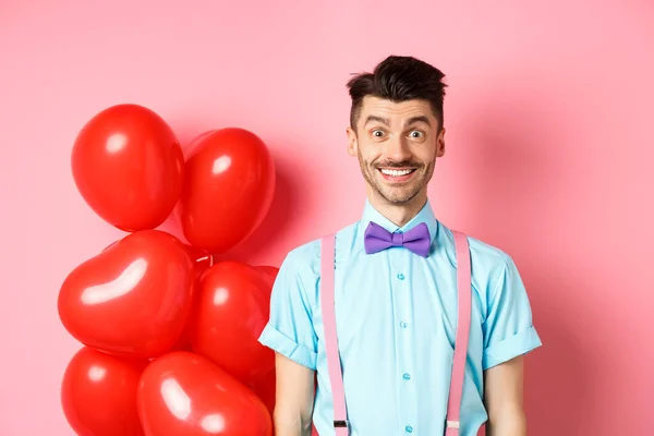 Concepto de San Valentín. Imagen de un joven guapo que se ve emocionado y sorprendido, sonriendo mientras está de pie sobre un fondo rosa romántico cerca de globos del corazón —  Fotos de Stock