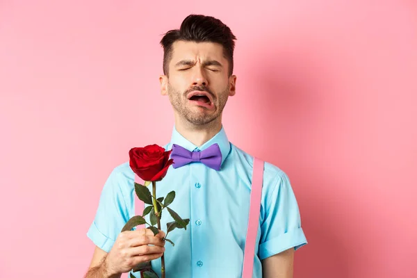 Chico desconsolado en divertida pajarita llorando por su novia, de pie solo con rosa roja sobre fondo rosa y sollozando, ruptura en el día de San Valentín —  Fotos de Stock